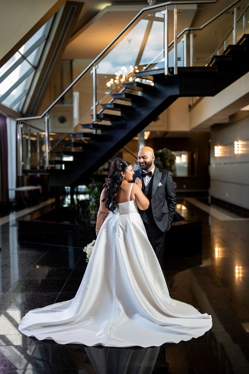 A couple in wedding attire share an intimate moment on a polished floor beneath a modern staircase with dramatic lighting.