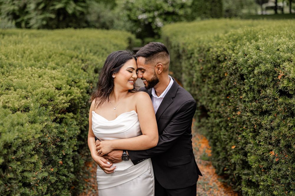 A couple embraces romantically in a lush green hedge maze, bride in white gown and groom in black suit.
