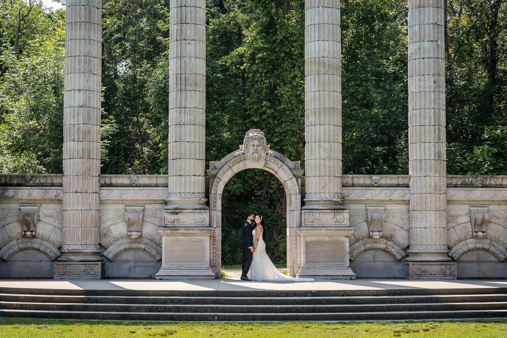 Elegant wedding couple poses between classic stone columns in a formal garden setting.