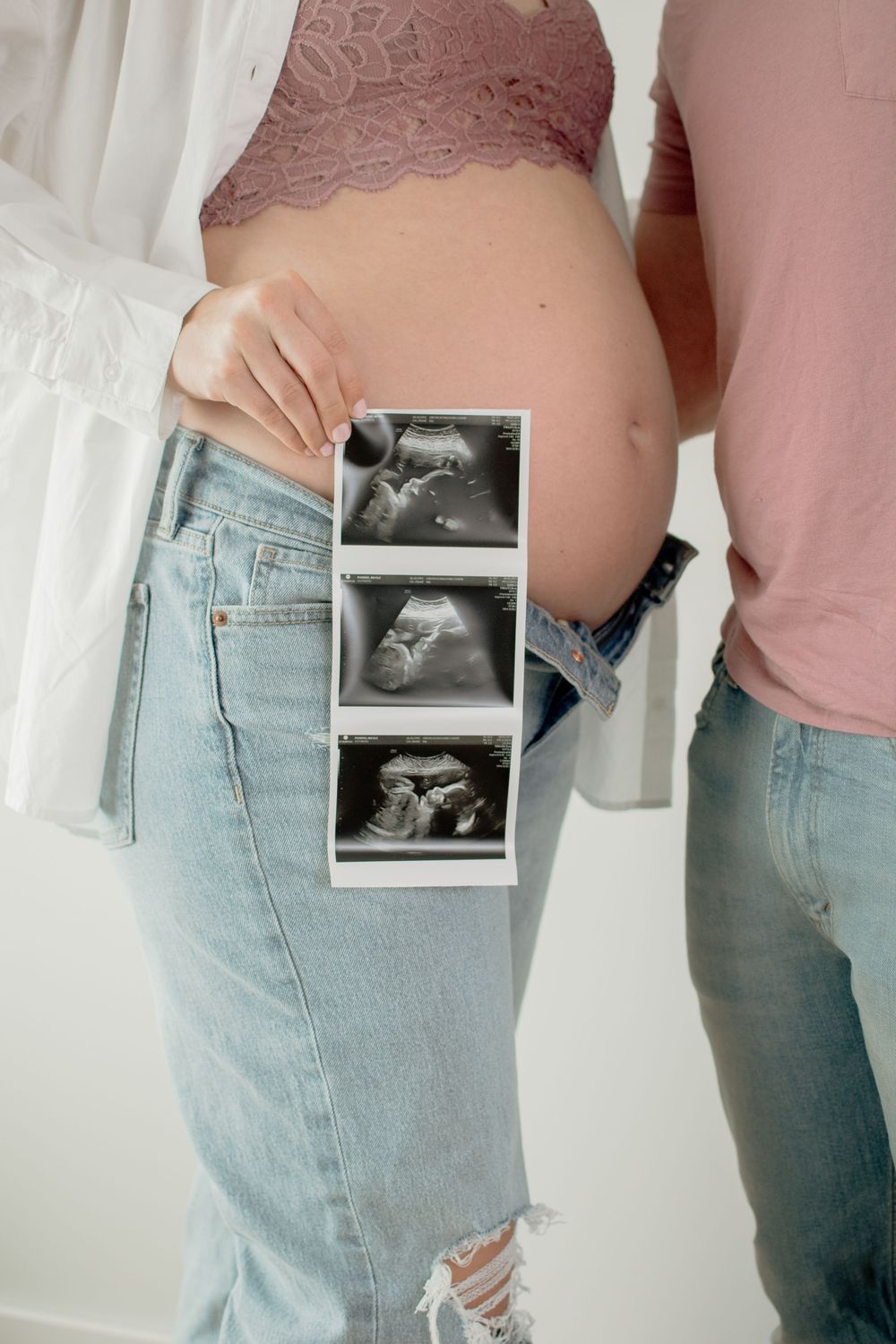 Pregnancy progress photos showing baby ultrasound held against denim pocket while wearing a pink top and light jeans.