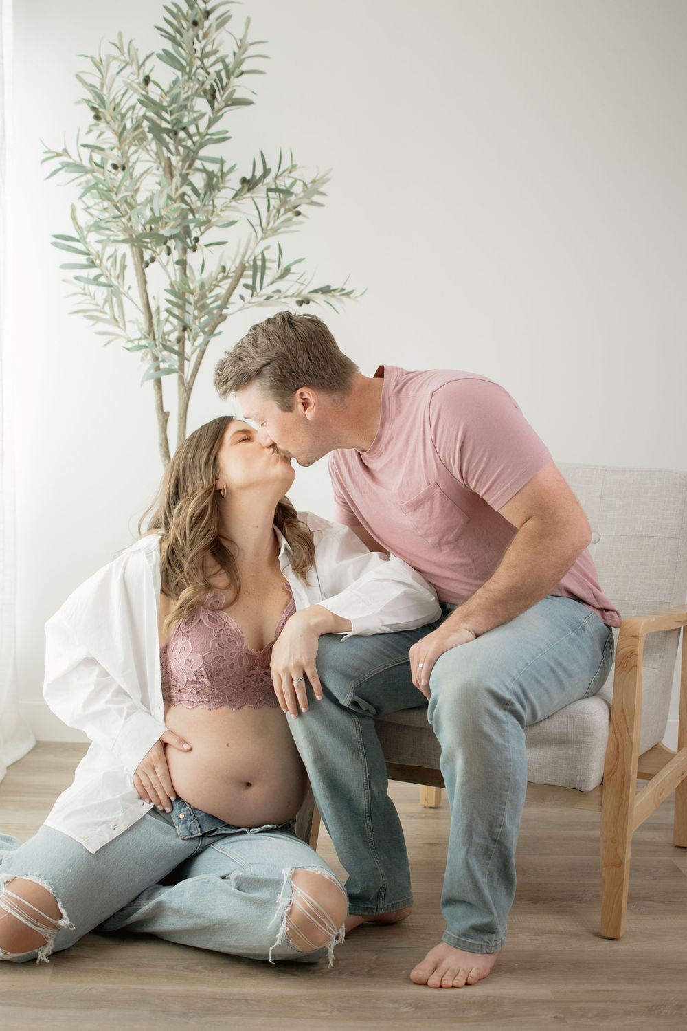 Intimate maternity photo session featuring couple in casual pink and blue outfits posing beside potted olive tree.