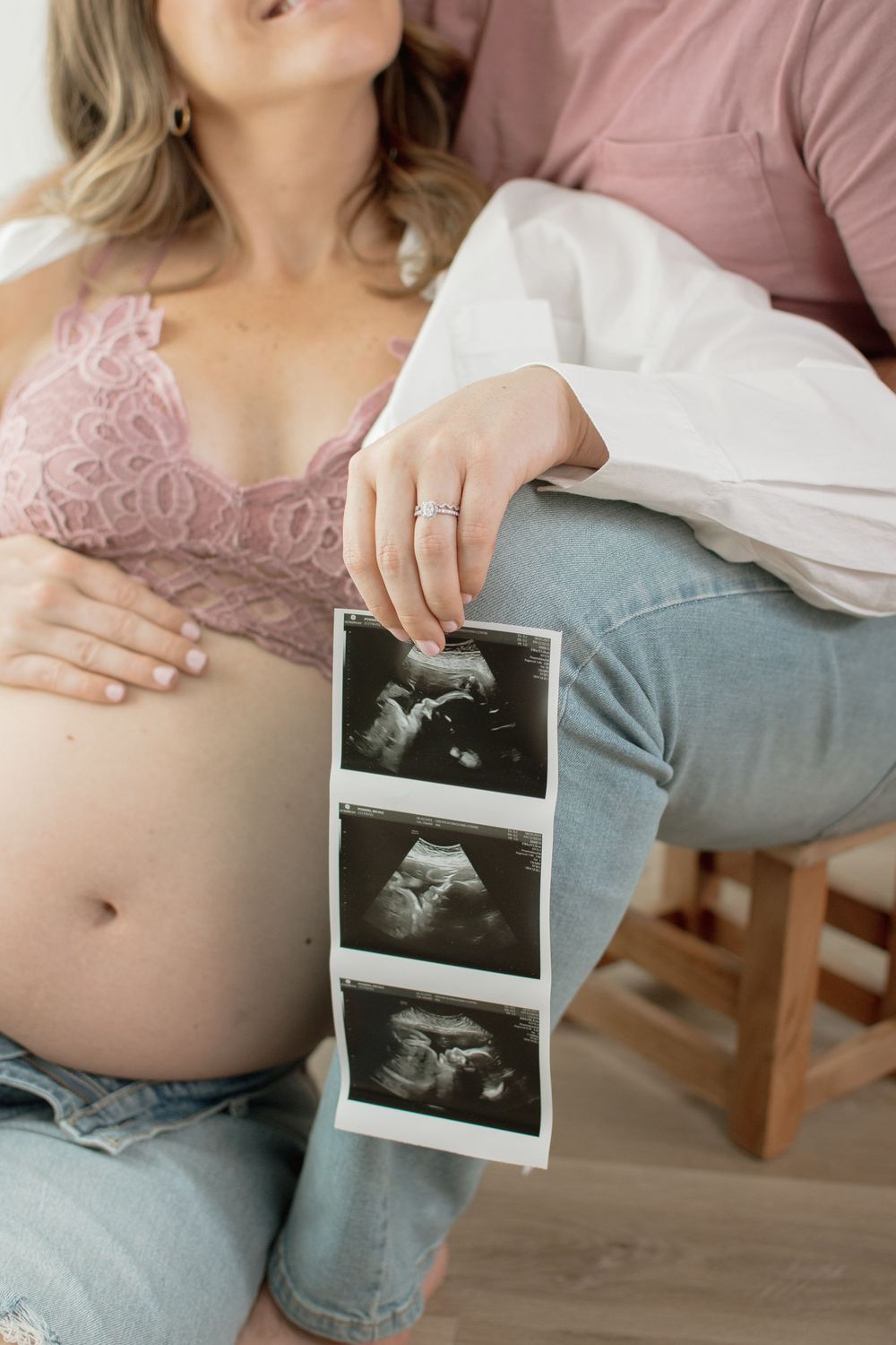 A pregnant person in jeans holds ultrasound photos while sitting on a couch, showing a growing baby bump.