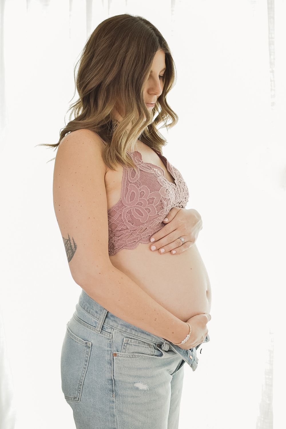 A pregnant woman in a pink bralette and light jeans poses for a maternity photo series against a white background.