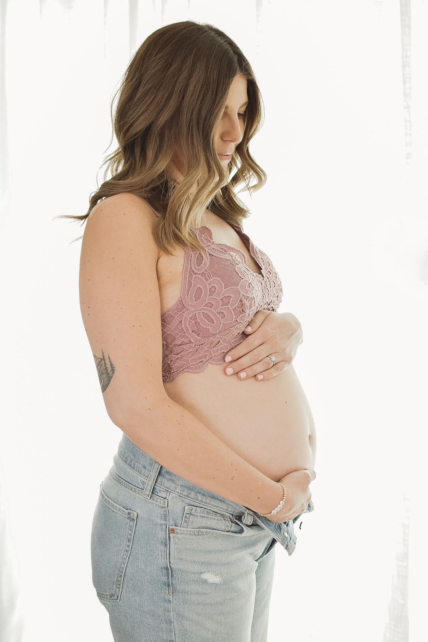 A pregnant woman in a pink bralette and light jeans poses for a maternity photo series against a white background.