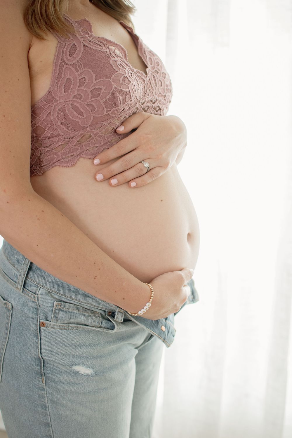 Multiple shots of a pregnant woman's belly shown in progression while wearing a pink lace top and jeans against a white backdrop.