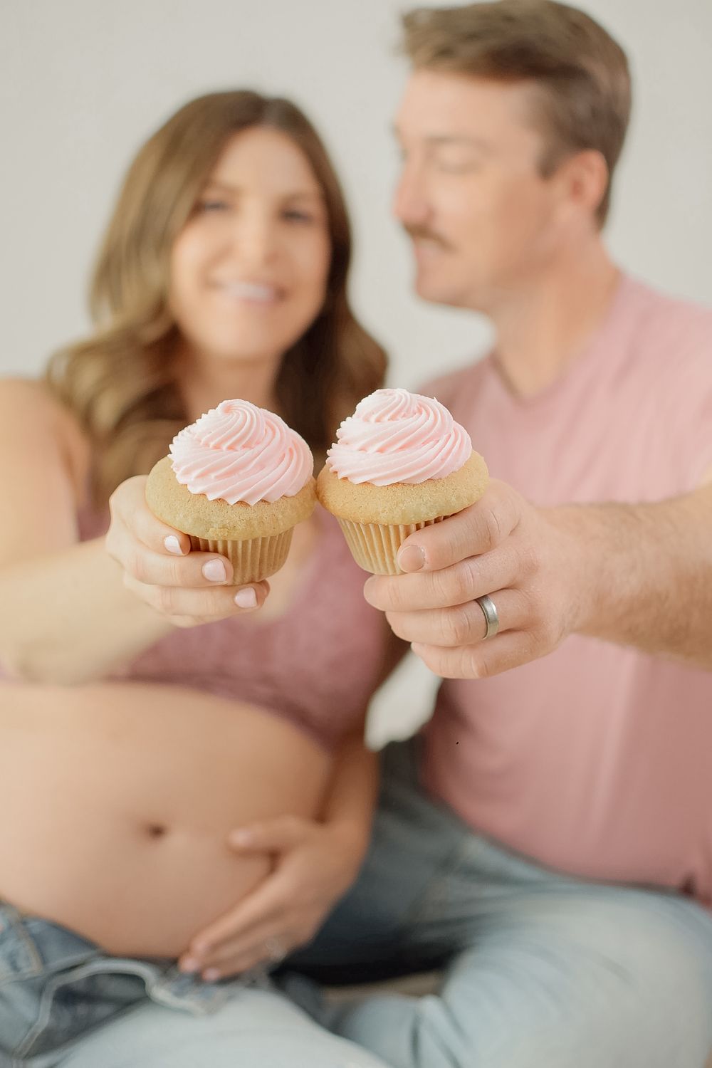 A couple in matching pink outfits holds frosted cupcakes while sitting together in a cozy indoor setting.