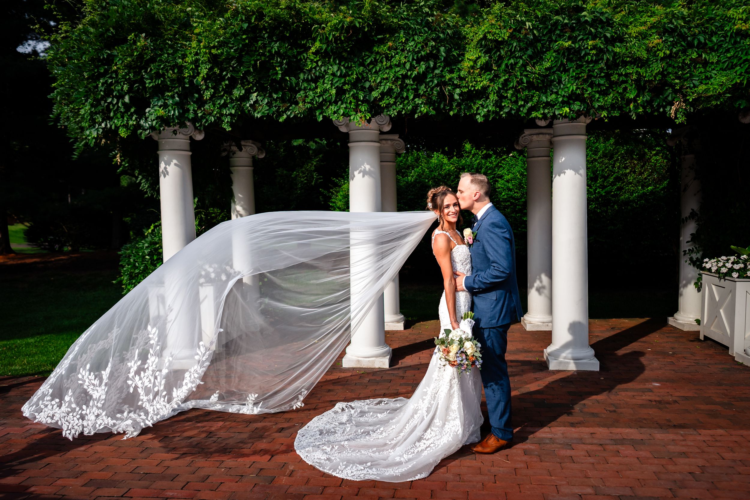 Wedding couple embraces under white columns as bride's lace veil flows dramatically in the wind.