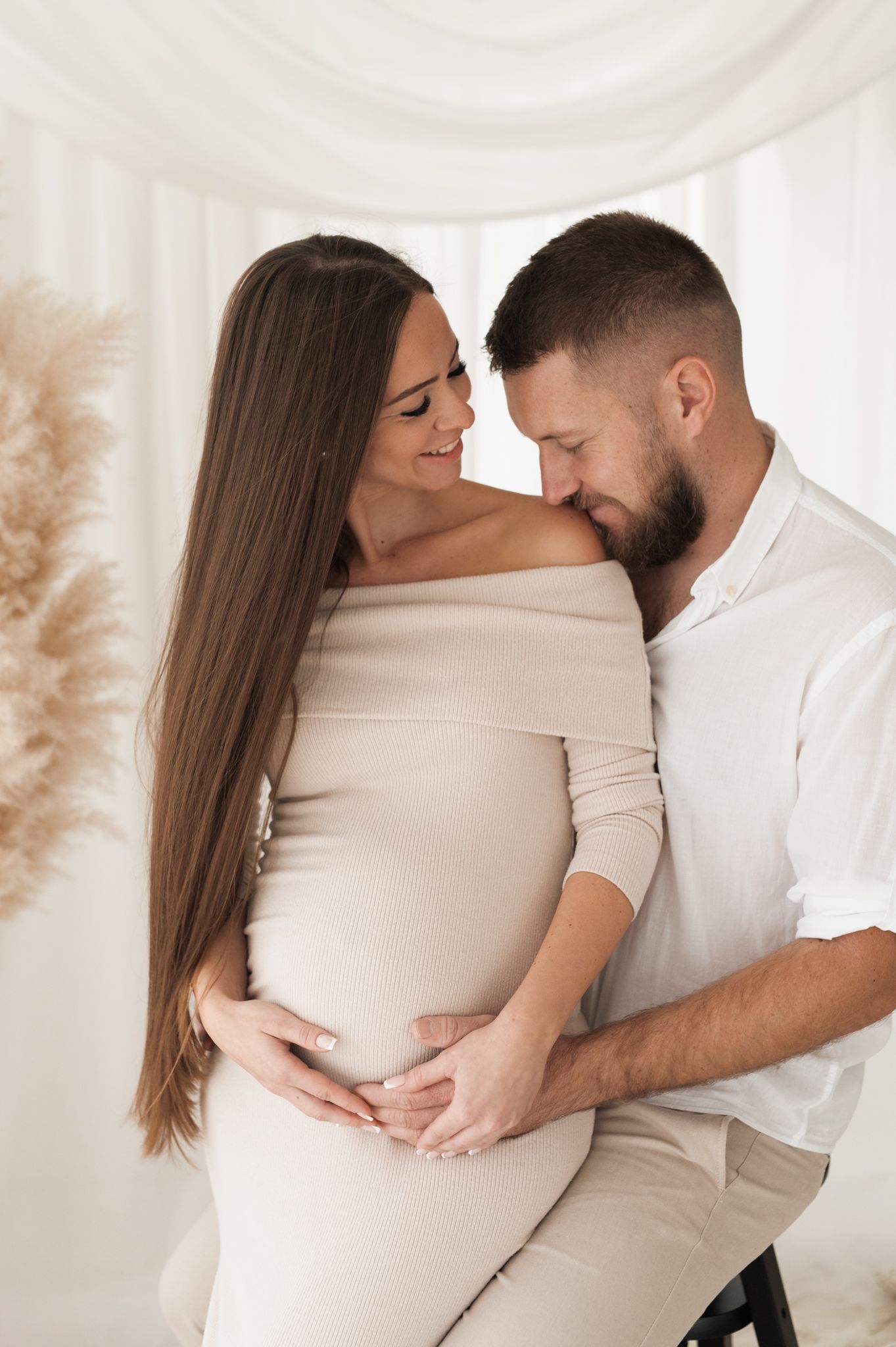 Fotografija para v studiu – nosečnica v svetli rebrasti obleki sedi v objemu partnerja, med njima nežnost in pričakovanje novega živlejnja.