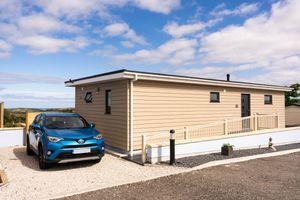 Modern beige mobile home with a blue hybrid car parked in the driveway on a sunny day with white clouds in the sky.