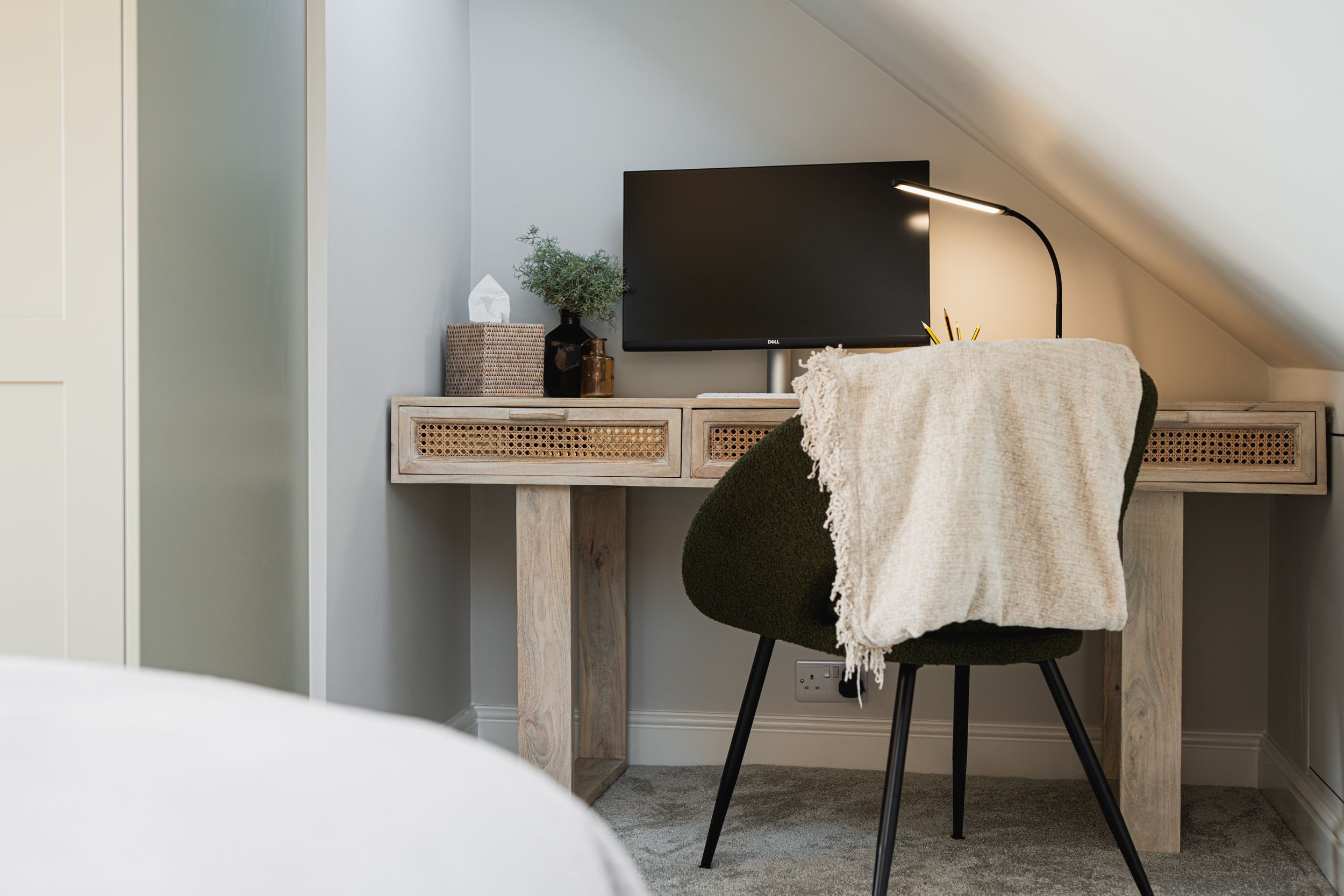 Minimalist home office setup with wooden desk, black chair, and monitor against white walls under slanted ceiling.