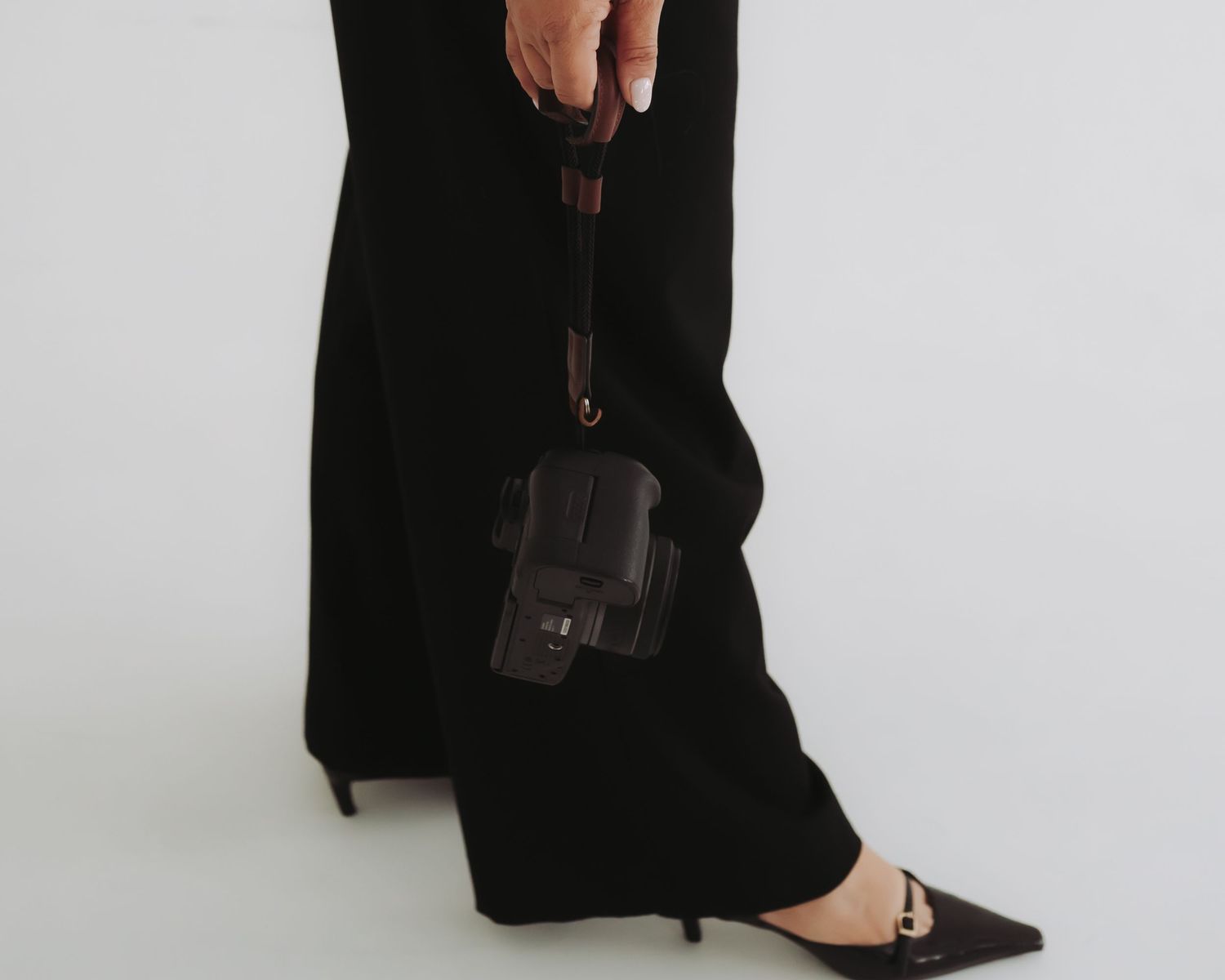 A person wearing black pants and pointed flats holds a small black leather handbag against a white background.