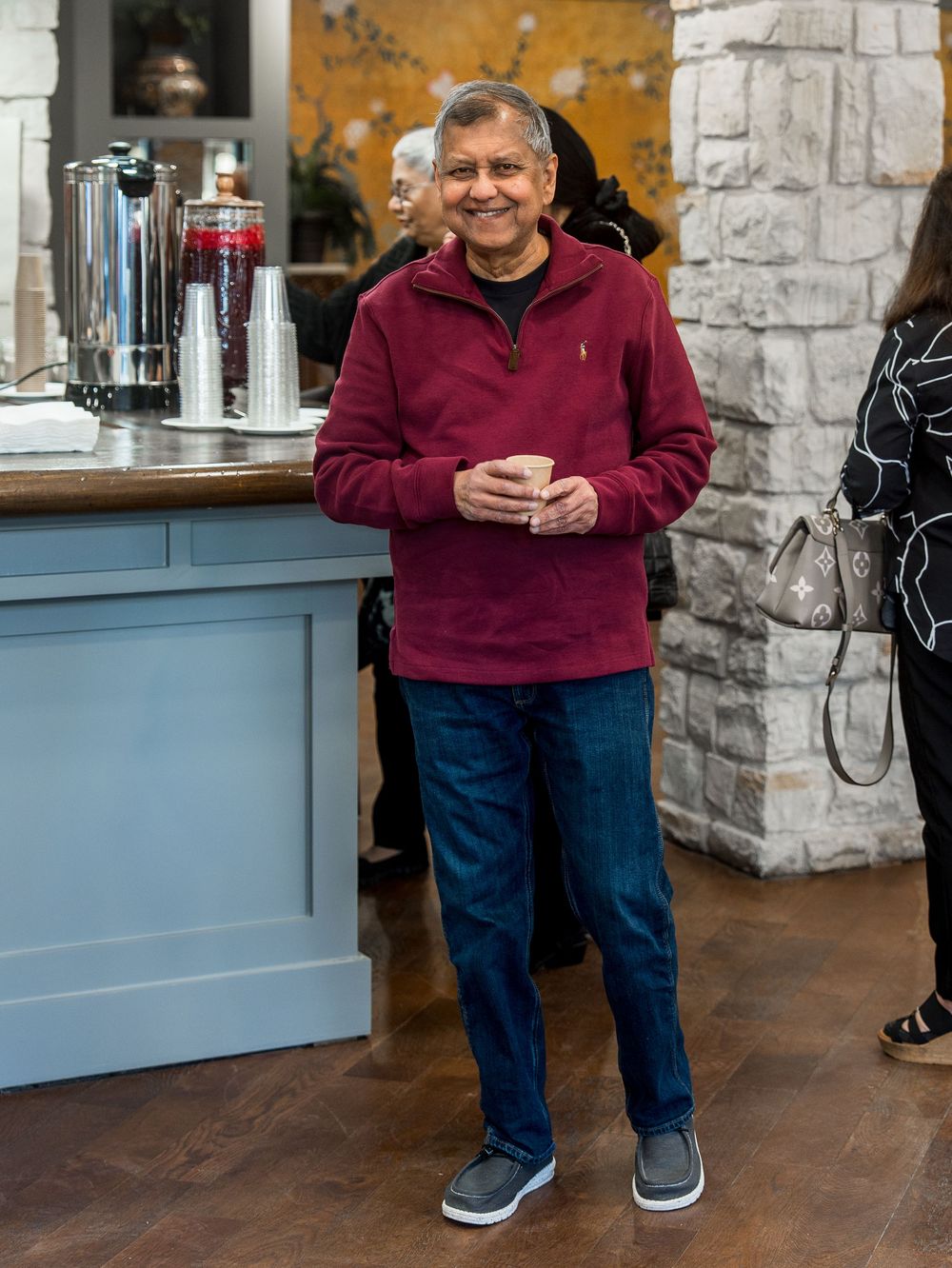 Person wearing a burgundy sweater and blue jeans looks at phone while standing in a room with stone walls.