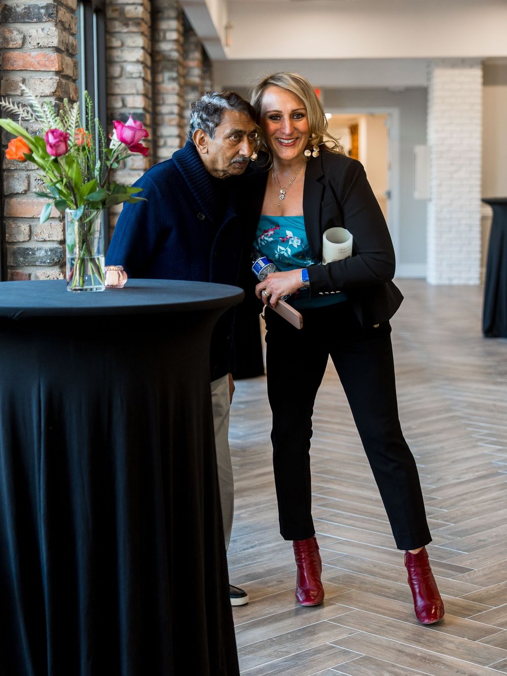 Two people in black attire stand together on hardwood flooring with bright red footwear visible.