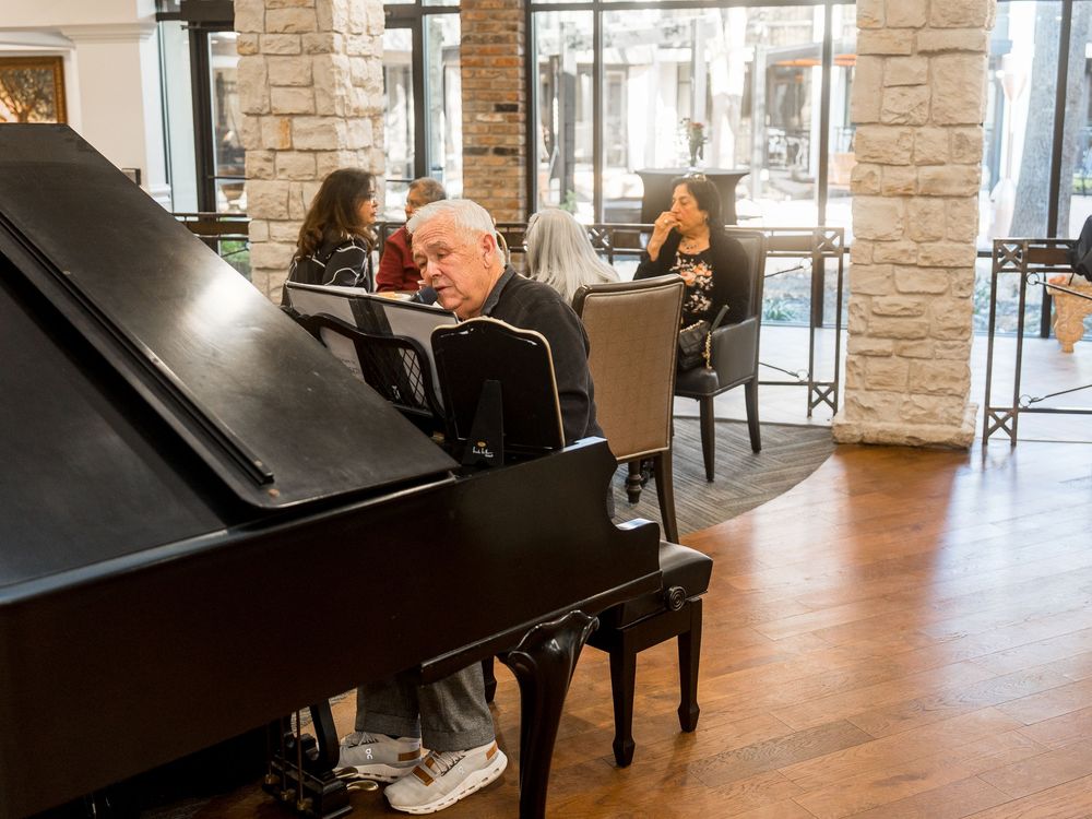 Senior sitting at a piano keyboard in a spacious room with stone walls and natural lighting.