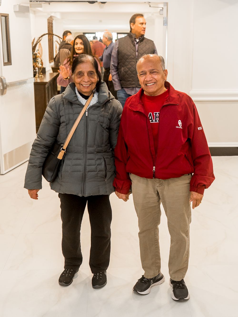 Two people walk together in a white hallway wearing winter jackets and casual attire.