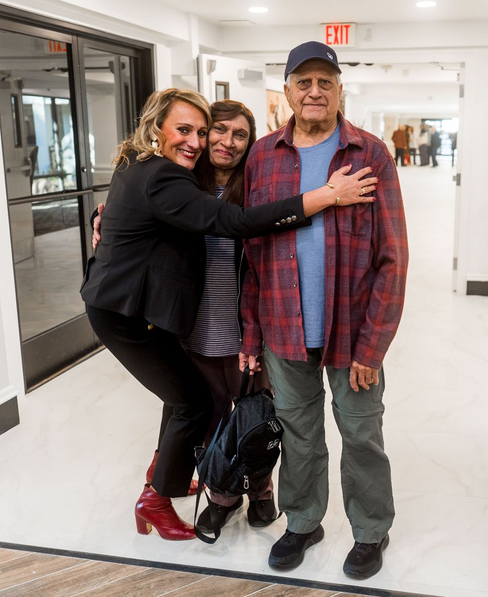 Three people pose together for a photo in a bright white hallway with an exit sign visible.