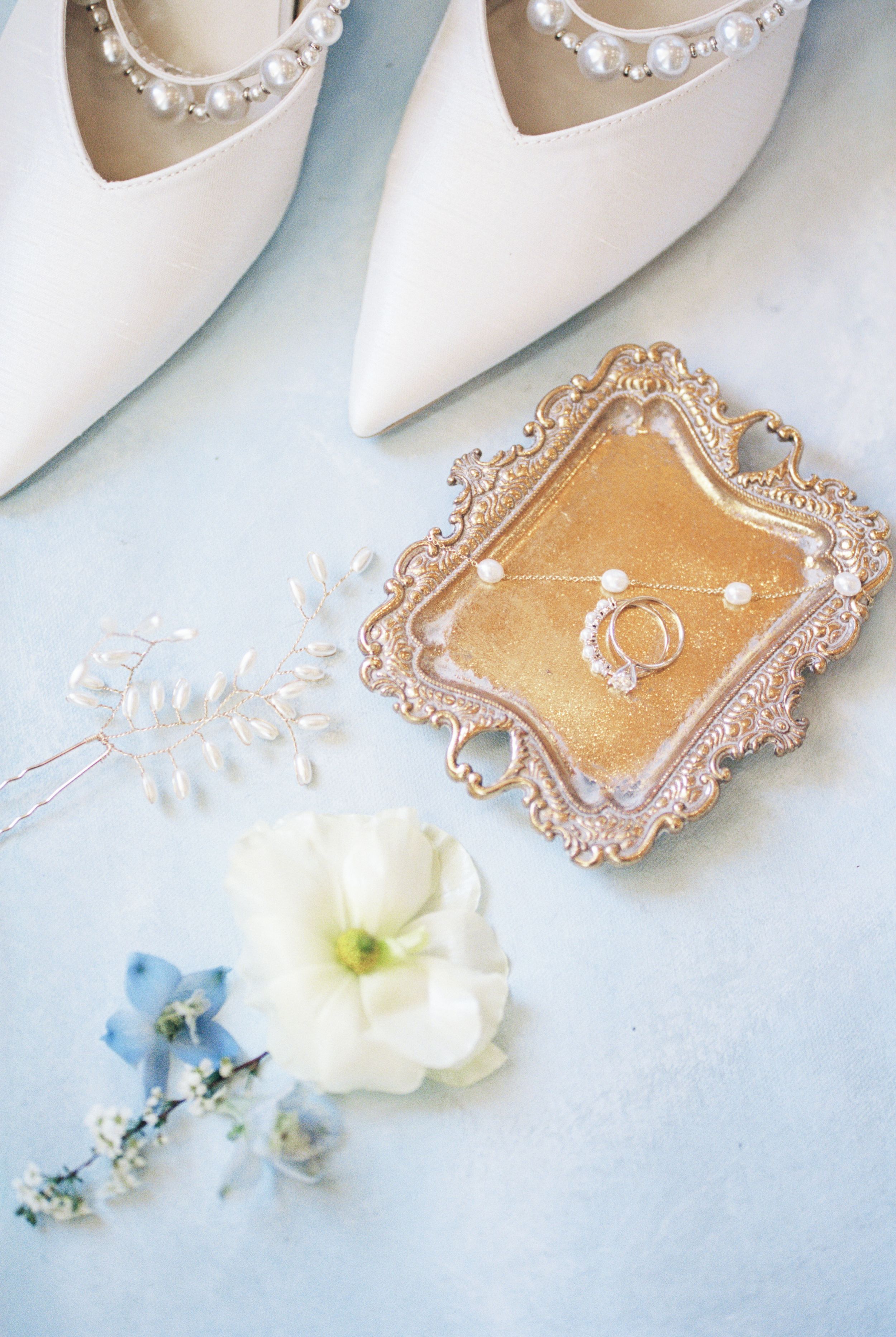 White wedding shoes with pearl embellishments are arranged with delicate blue flowers and petals on a light surface.