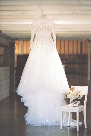 An elegant white lace wedding dress with long sleeves hangs beside a small white chair with a bouquet.