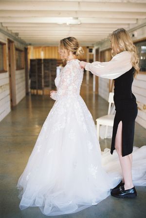 A woman in a black dress helps adjust a bride's lace wedding gown in an indoor hallway.