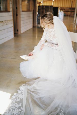 A bride in a long-sleeved lace gown adjusts her flowing tulle dress on a polished floor.