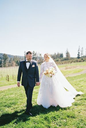 A newlywed couple walks hand in hand through a sunny meadow with mountains in the background.