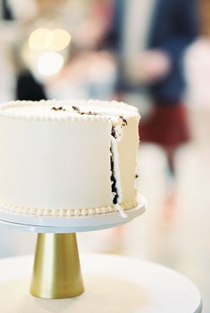 A simple white wedding cake with minimal decoration displayed on a gold pedestal stand.
