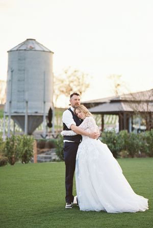 Couple embraces on manicured lawn with rustic silo in background during sunset.