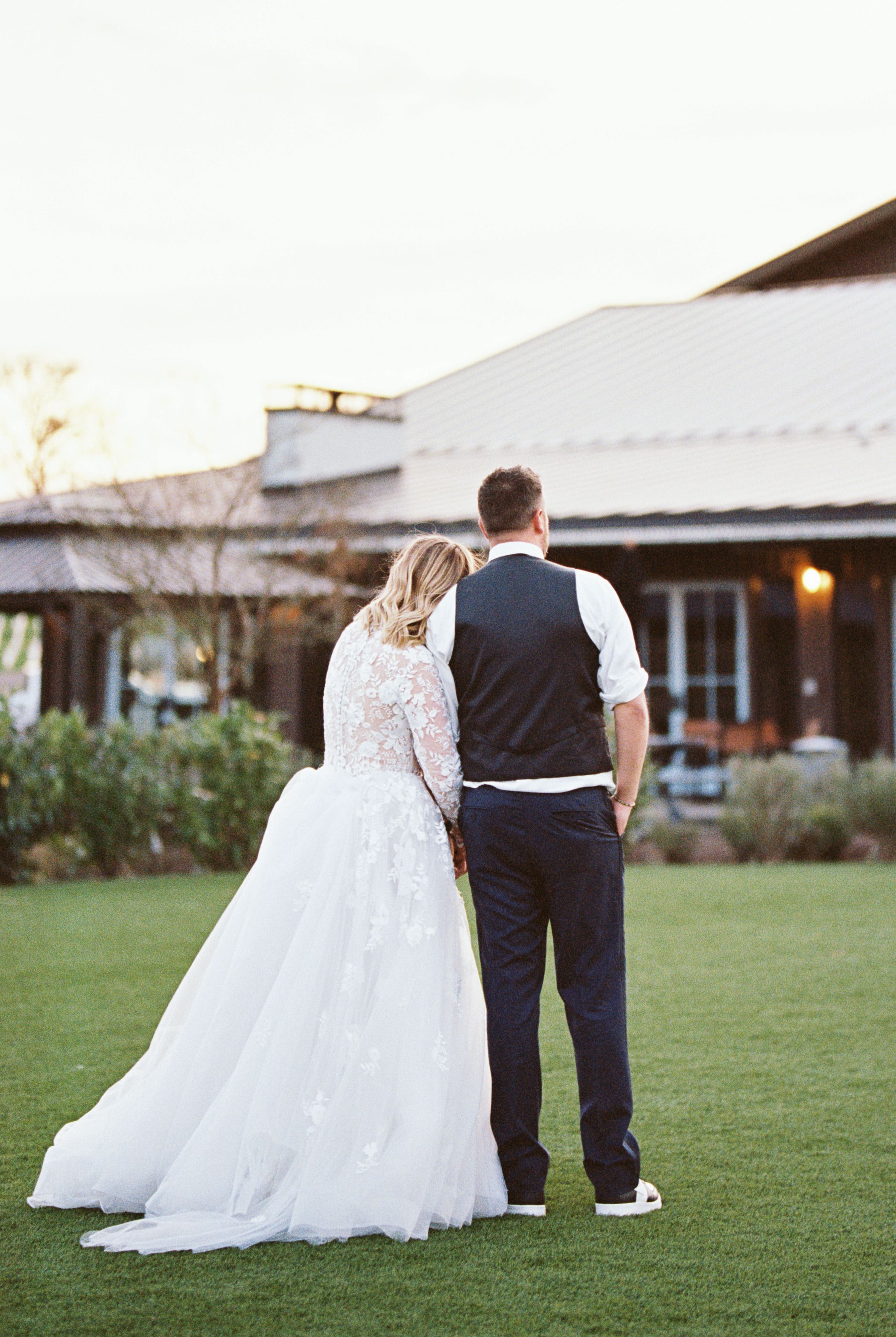 Romantic back view of couple in wedding attire standing on green lawn at dusk.