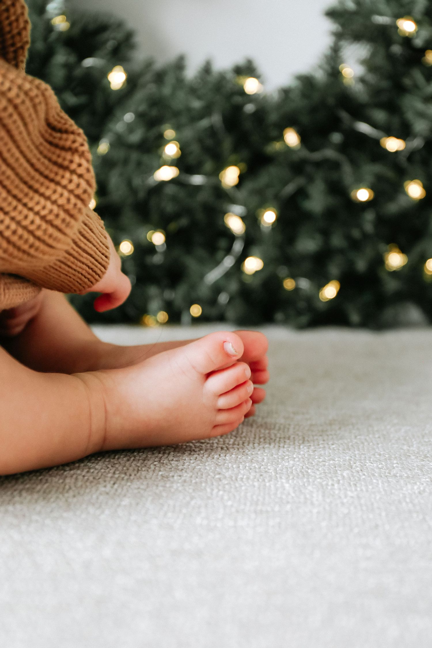 Small barefoot sitting on white carpet with soft Christmas wreath lights twinkling in bokeh background during photoshoot  in Nottingham