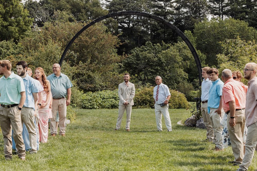 Wedding party members in pastel outfits gather for group photos on a lush green lawn surrounded by trees.