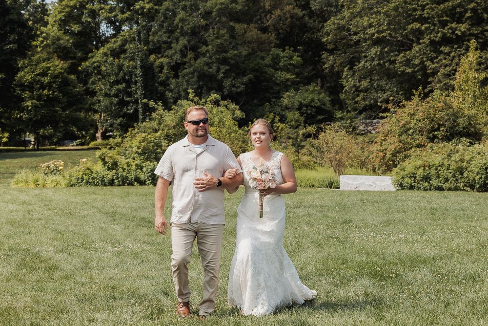 A couple walks together on a grassy lawn surrounded by lush trees on a sunny summer day.