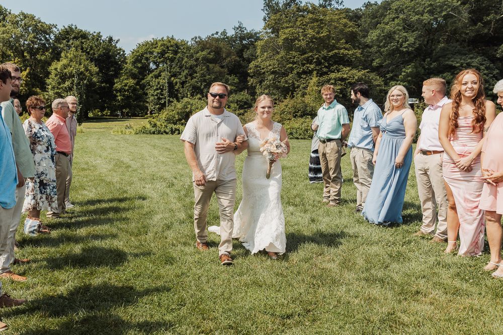 People in colorful dresses and suits gather outdoors on a grassy hillside with trees on a sunny day.
