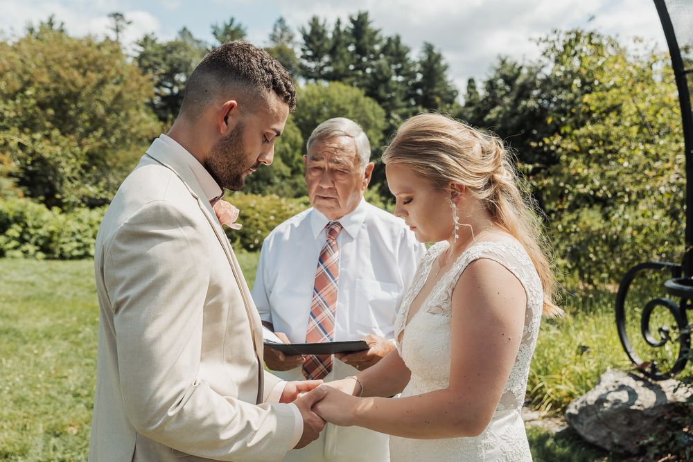 An outdoor wedding ceremony takes place in a garden setting with greenery in the background.