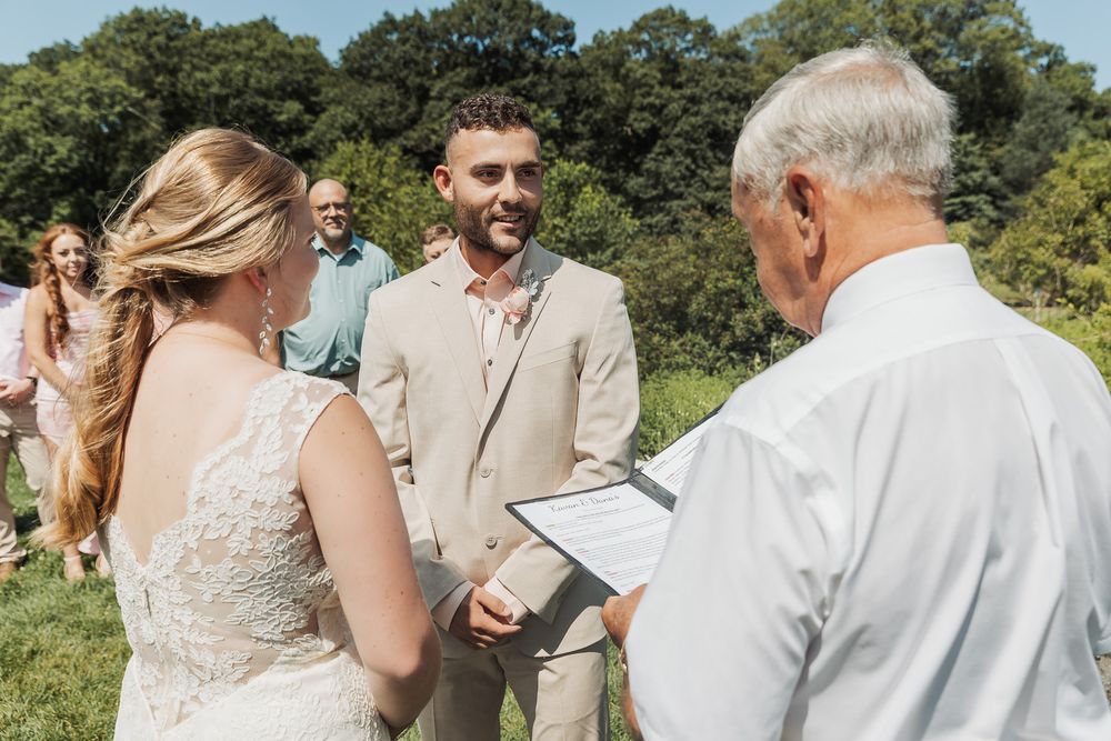 Wedding ceremony taking place outdoors on a sunny day with guests gathered in a garden setting.