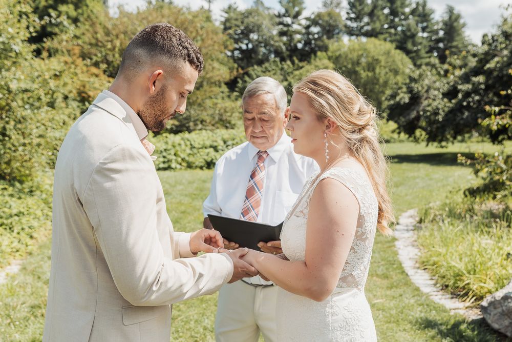 A couple shares an intimate moment exchanging rings during an outdoor summer wedding ceremony in a lush garden setting.