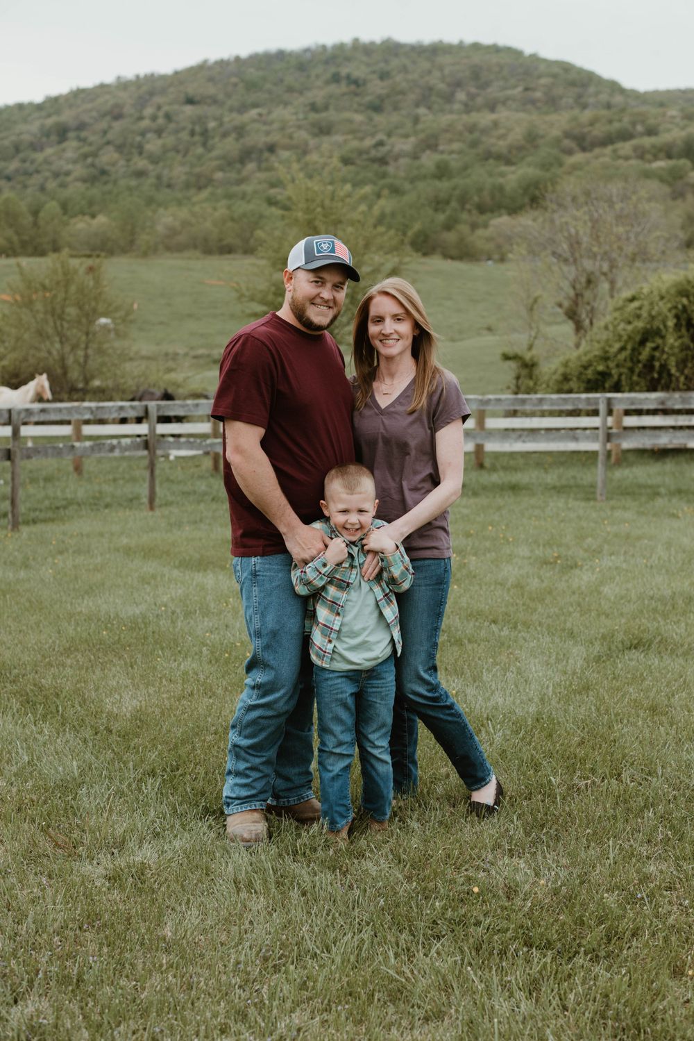 Casual family portrait session in a grassy field with wooden fencing and rolling hills.
