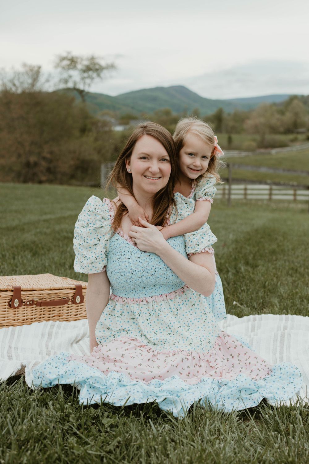 A tender outdoor photoshoot featuring floral dresses against a pastoral mountain backdrop with a picnic blanket.