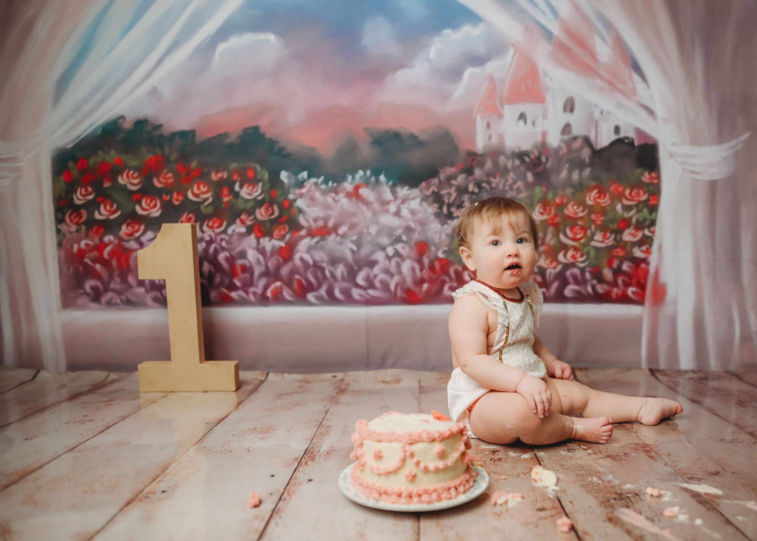 First birthday celebration setup with cake against a painted floral backdrop on wooden floor.