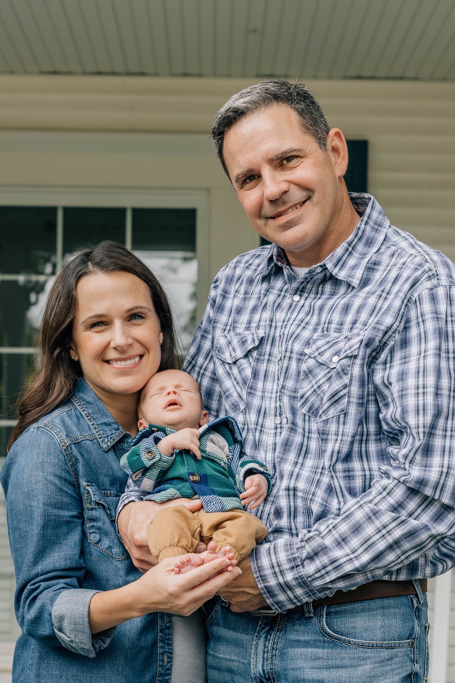 Smiling couple holding their newborn baby in front of their home for a family portrait photo session.