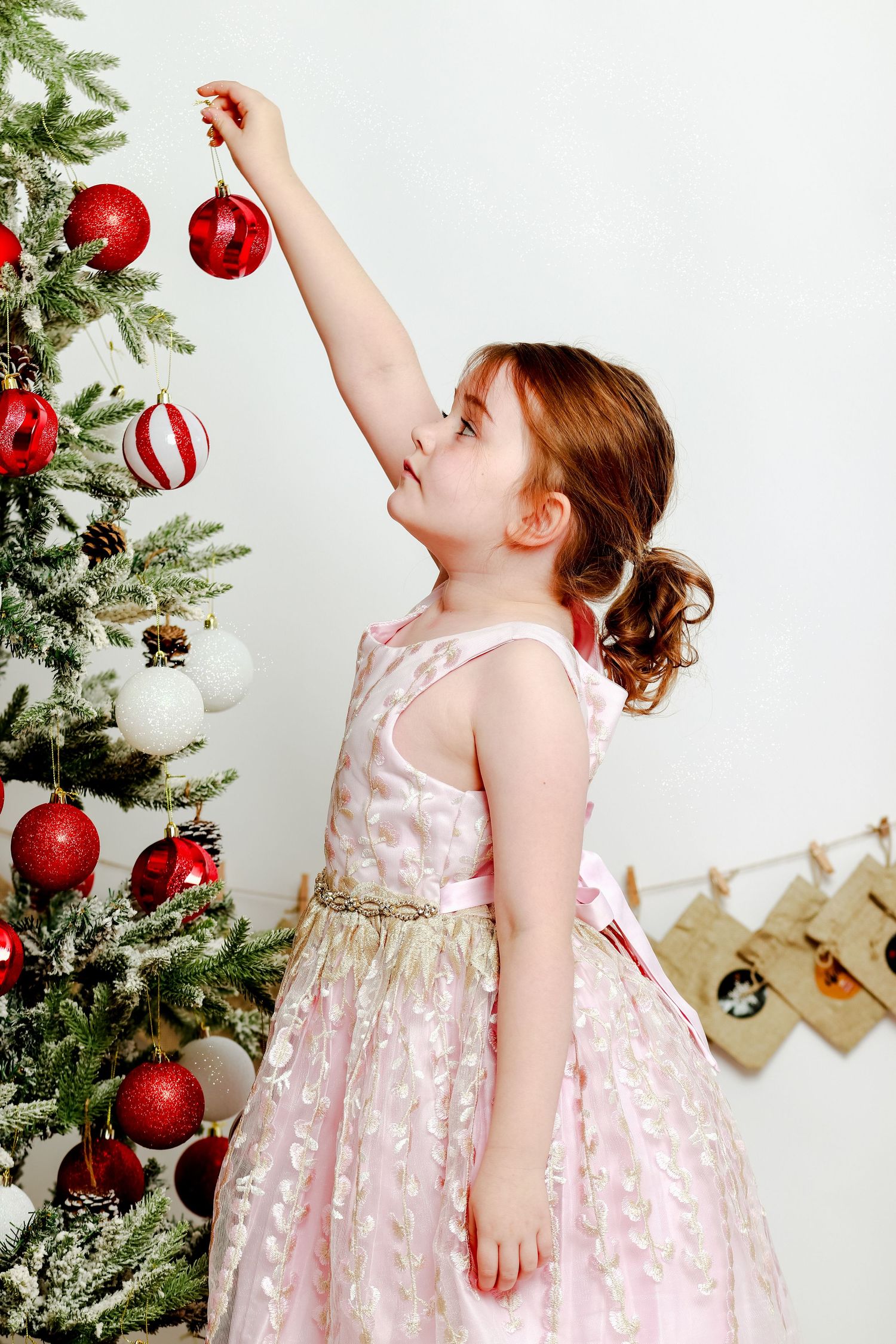 Child in pink dress decorating Christmas tree with red ornaments.