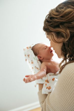 Mother in cream sweater holds and kisses newborn baby in white outfit against neutral background.