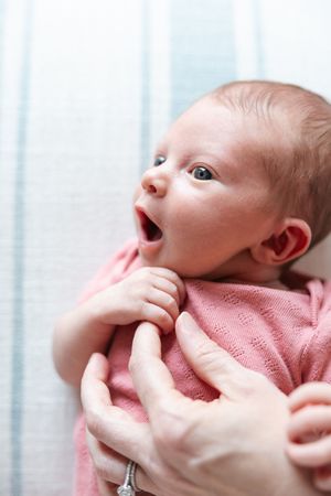 A tiny baby in a pink outfit looks up with wide eyes and an open mouth while being held by gentle hands.