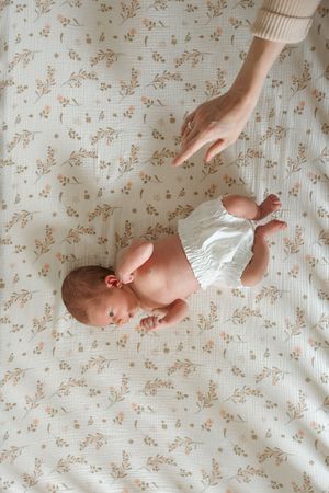 A gentle hand reaches toward a newborn baby lying on a floral patterned blanket wearing a white diaper cover.