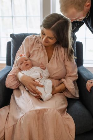 Family moment on grey couch with baby in white outfit and parent in pink dress.