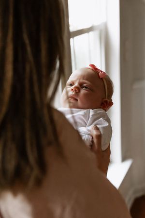 Baby wearing white clothing and red headband looks up while being held near a window.
