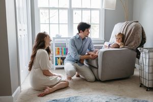 Three people gather around a cozy gray armchair in a bright room with window light and built-in bookshelves.