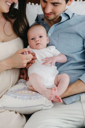 A tender moment with a baby in white onesie nestled between adults on a bed.