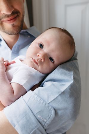 A baby with bright eyes rests on someone's shoulder wearing a light blue shirt.