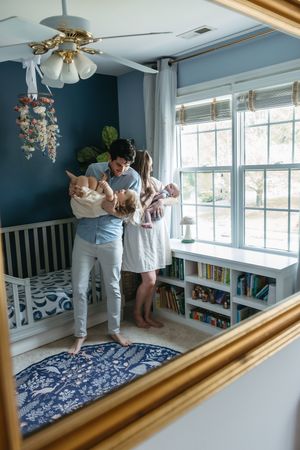 A mirror reflection shows a nursery with navy walls, white furniture, and a decorative crystal chandelier near a sunny window.