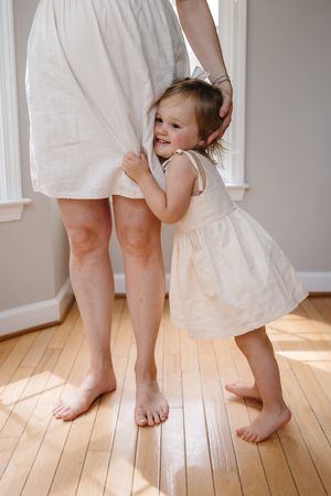 A small child in a white dress hugs an adult's legs while standing on hardwood flooring in a sunlit room.